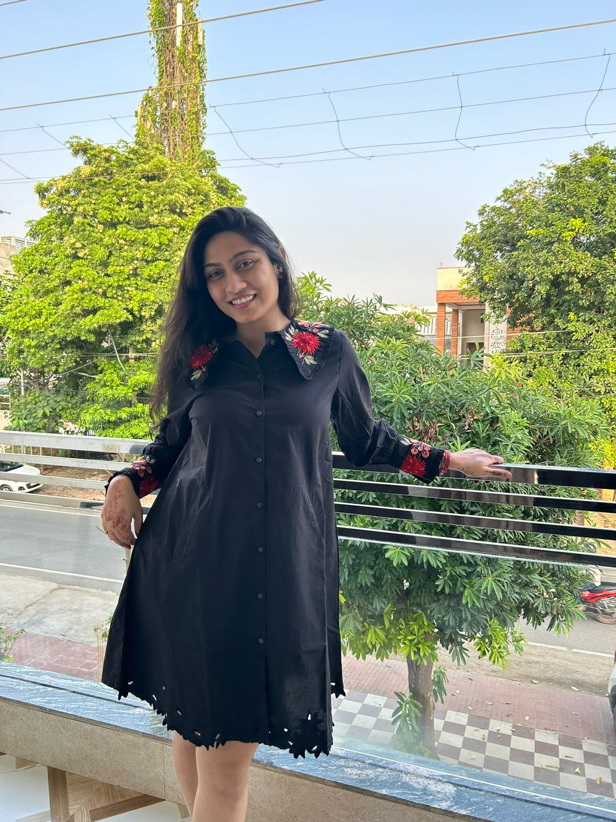 Woman in a black dress with floral embroidery standing outdoors on a sunny day.