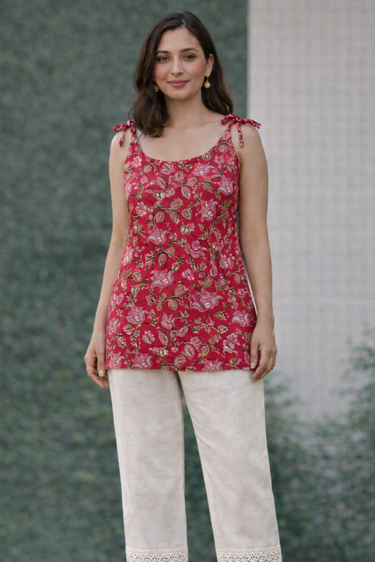 Woman wearing a red floral sleeveless top and white pants with lace trim against a green wall.