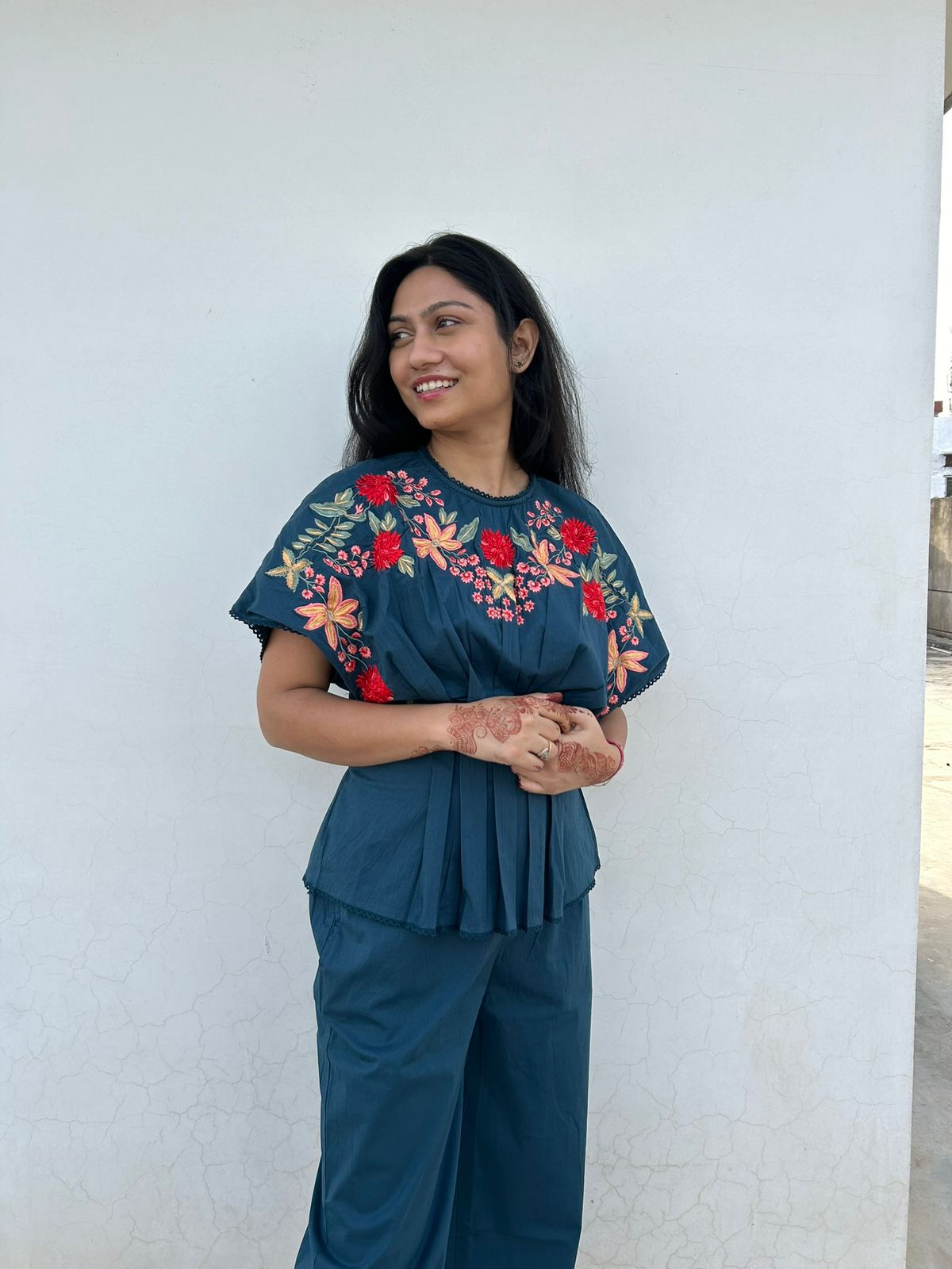 Woman wearing a teal outfit with floral embroidery against a white wall