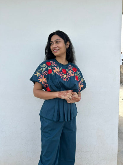 Woman wearing a teal outfit with floral embroidery against a white wall