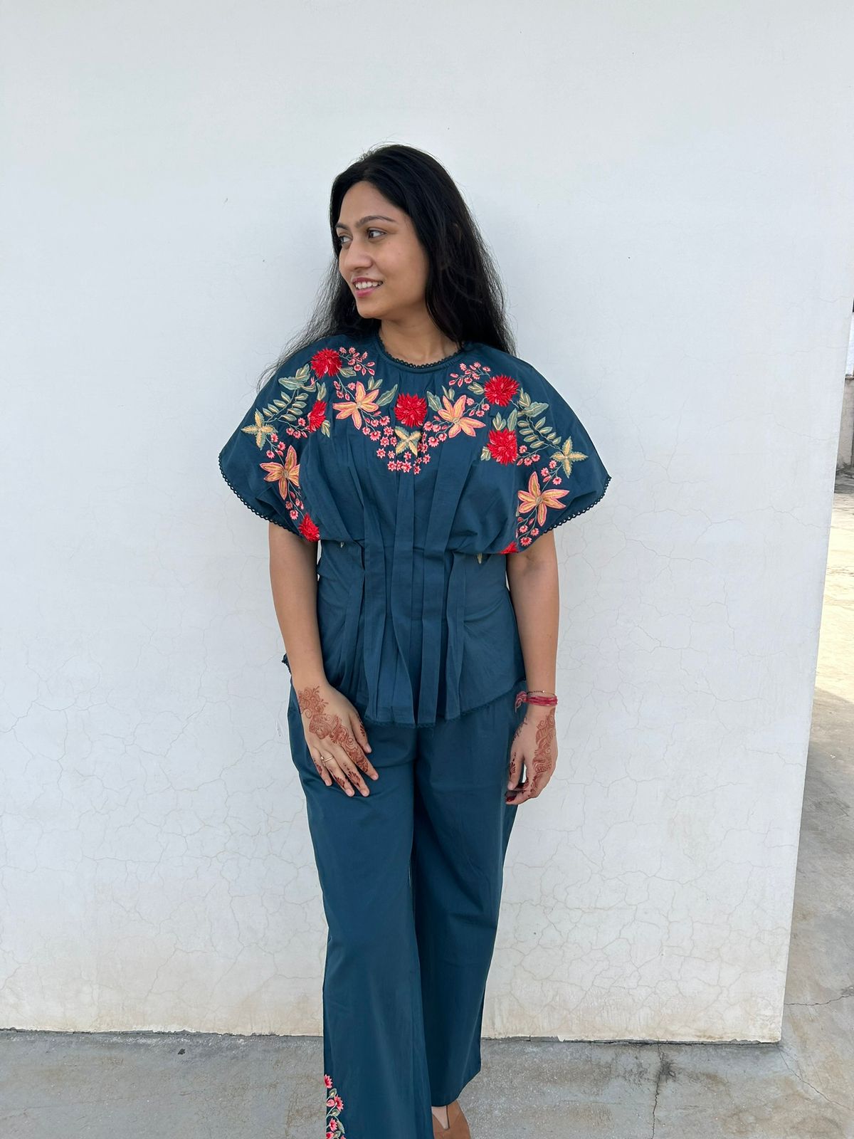 Woman wearing a teal outfit with floral embroidery against a white wall.